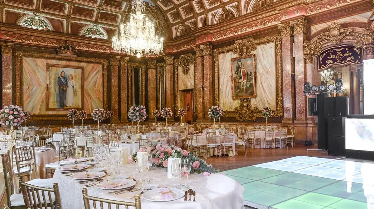 Interior del Salón de los Reyes en el Casino Español con mesas, candelabros y pinturas. Se ven centros de mesas con flores. Se ve una pista de baile.